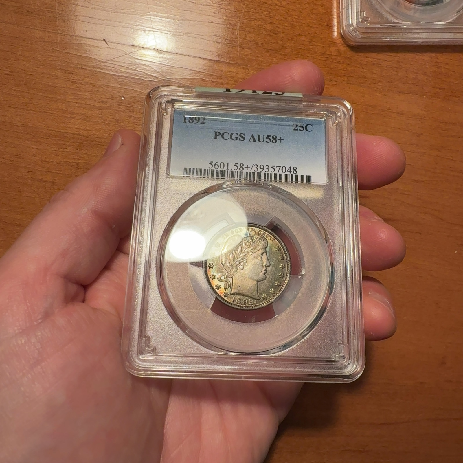 Handheld phone photograph of a 1892 quarter · PCGS AU58+ in its holder, with glare and an everyday background